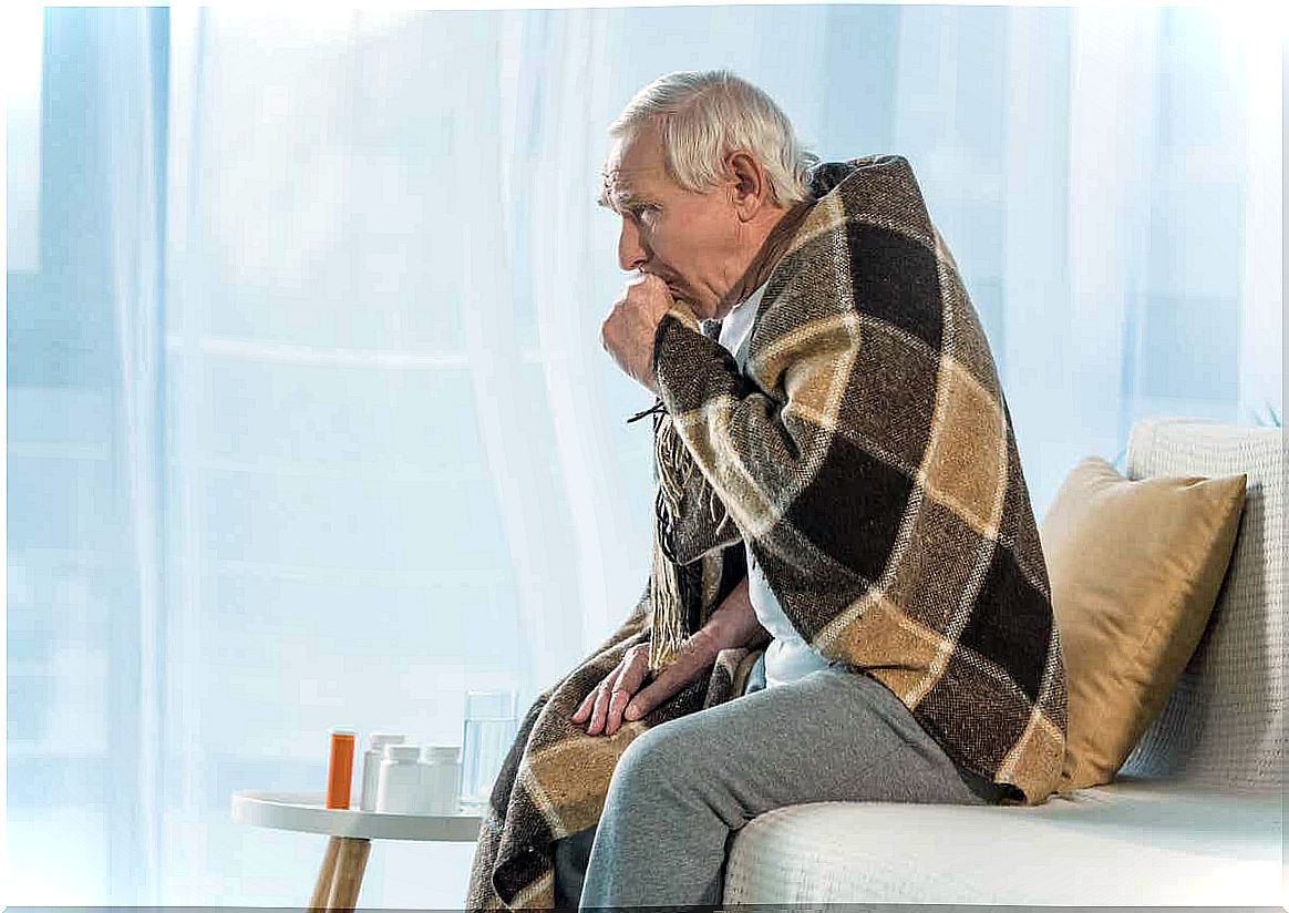 An elderly man with a blanket over his shoulders coughs in his hand